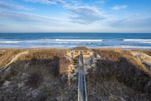 A beachside walkway leading to a wooden structure with a thatched roof, surrounded by sand and dry grass. - 5691- Michelitch - Oceanfront Nags Head Condo With 2 Balconies (Nags Head)