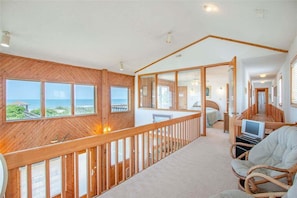 Ein geräumiger Raum mit Holzboden, großen Fenstern mit Meerblick, einem Balkon mit Geländer und einer Sitzecke mit Fernseher.