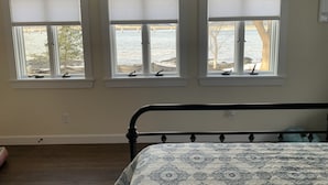 A bedroom with a bed, three windows with blinds, a view of the beach, and a wooden floor. - Cottage in Edgecomb - Waterfront Holiday Getaway (Edgecomb)