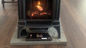 A wood-burning stove with a digital control panel, set against a brick wall and wooden floor, in a room with blue walls and a white chair. - Cottage in Edgecomb - Waterfront Holiday Getaway (Edgecomb)