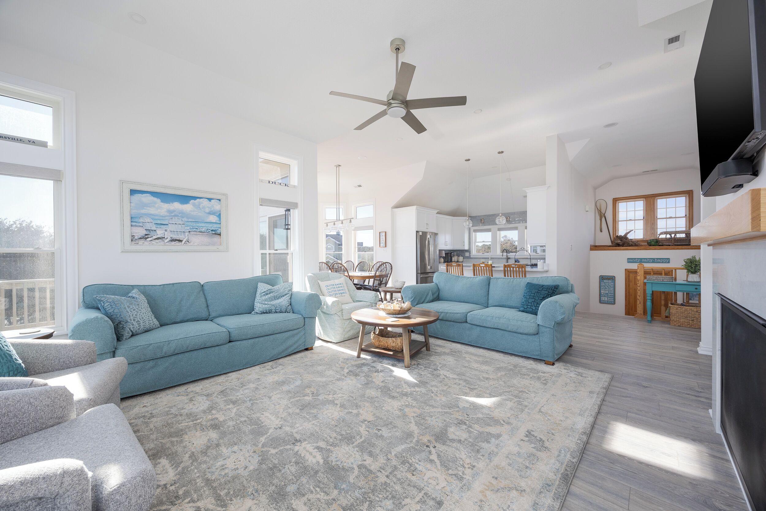 A spacious living area with two light blue sofas, a coffee table, and a large rug. There is a ceiling fan, a TV, and a view of the kitchen in the background.