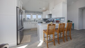 Een moderne keuken met witte kasten, een roestvrijstalen koelkast, een centraal kookeiland met barstoelen en uitzicht buiten door grote ramen.