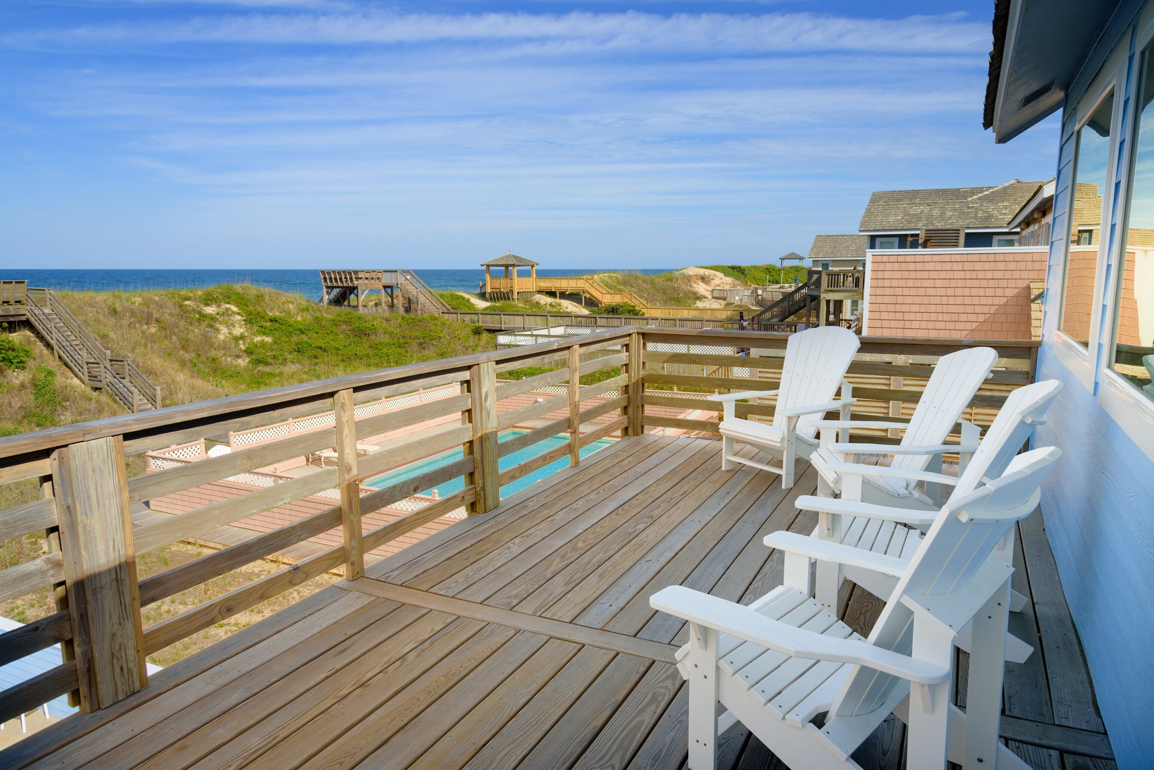 Terraza de madera con sillas blancas que da a una playa y una piscina.