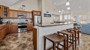 A spacious kitchen with wooden cabinets, stainless steel appliances, and a central island with bar stools. - 7700-Lee's Landing - Oceanfront Avon | Pool & Hot Tub | Sleeps 10 (Avon)