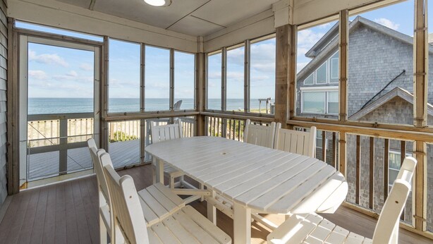 A covered outdoor area with a white table and benches, large windows, and a view of the ocean. - 7700-Lee's Landing - Oceanfront Avon | Pool & Hot Tub | Sleeps 10 (Avon)
