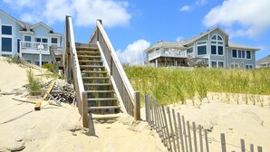 Ett hus vid stranden med en trappa som leder upp till en balkong, omgivet av sand och växtlighet.