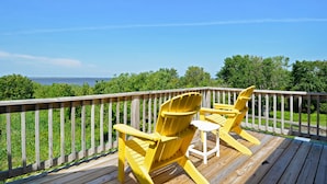 Eine Holzterrasse mit zwei gelben Adirondack-Stühlen und einem kleinen Tisch, mit Blick auf ein Gewässer und Grünflächen.