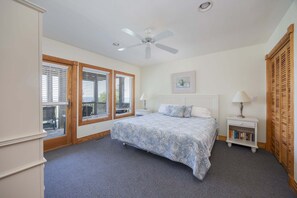 A bedroom with a bed, a ceiling fan, a window with blinds, a bedside table with a lamp, and a bookshelf.