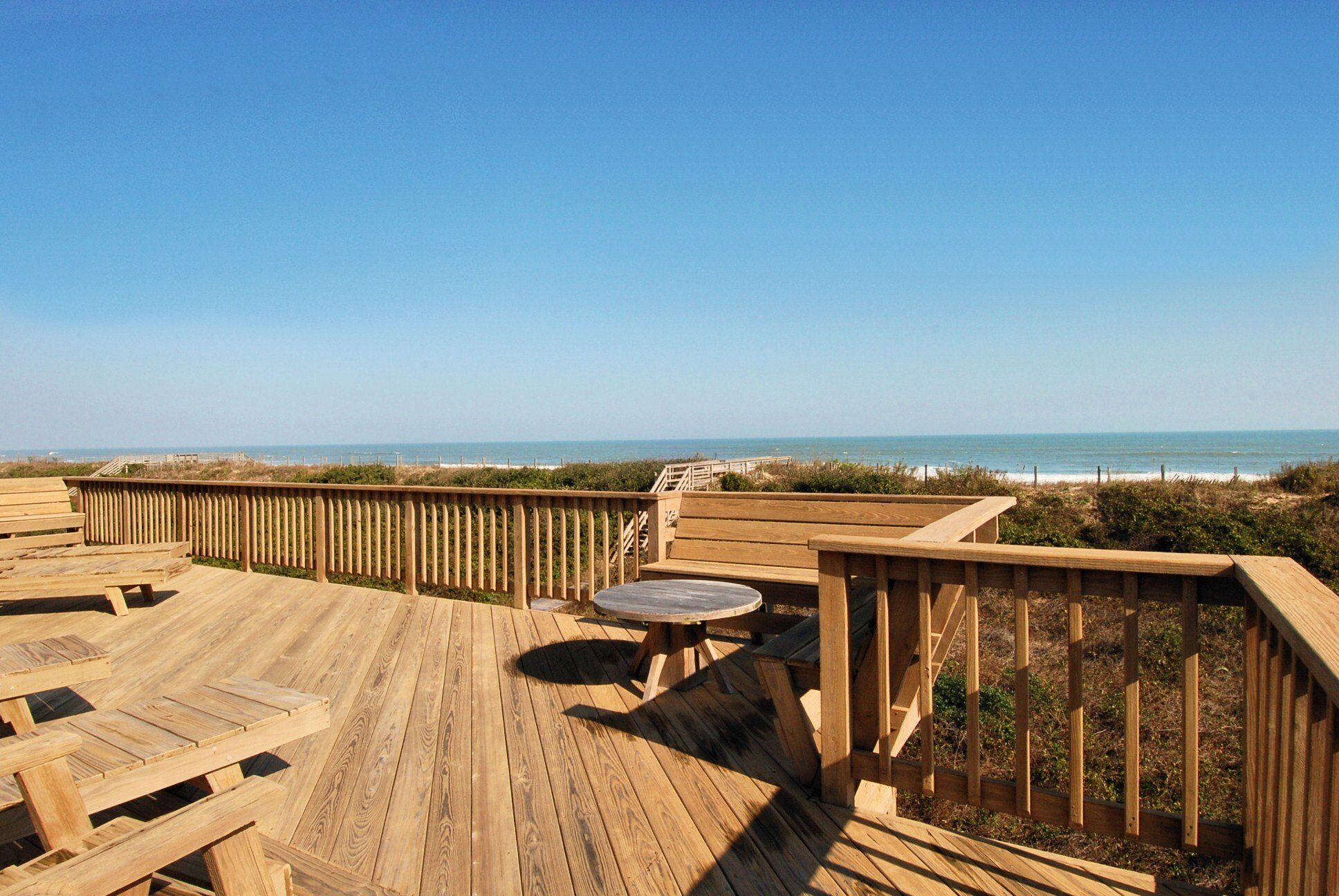 Una terraza de madera con una mesa y sillas, con vista a una playa y un cielo despejado.