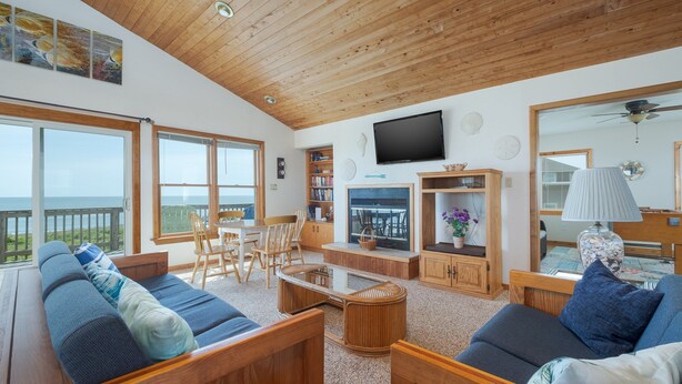 Amplia sala de estar con techo de madera, ventanas grandes, televisor y vista al océano.