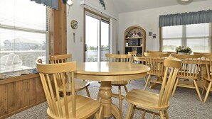Una habitación con una mesa redonda de madera y seis sillas de madera, una estantería con objetos decorativos, y una ventana con persianas.
