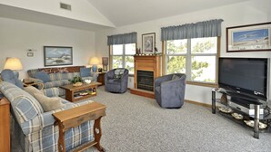Una sala de estar con un sofá, sillones, una chimenea, un televisor y ventanas grandes con cortinas.