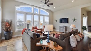 Un salon spacieux doté d’une grande fenêtre, d’un ventilateur de plafond et d’un coin salon confortable avec un canapé et des fauteuils.