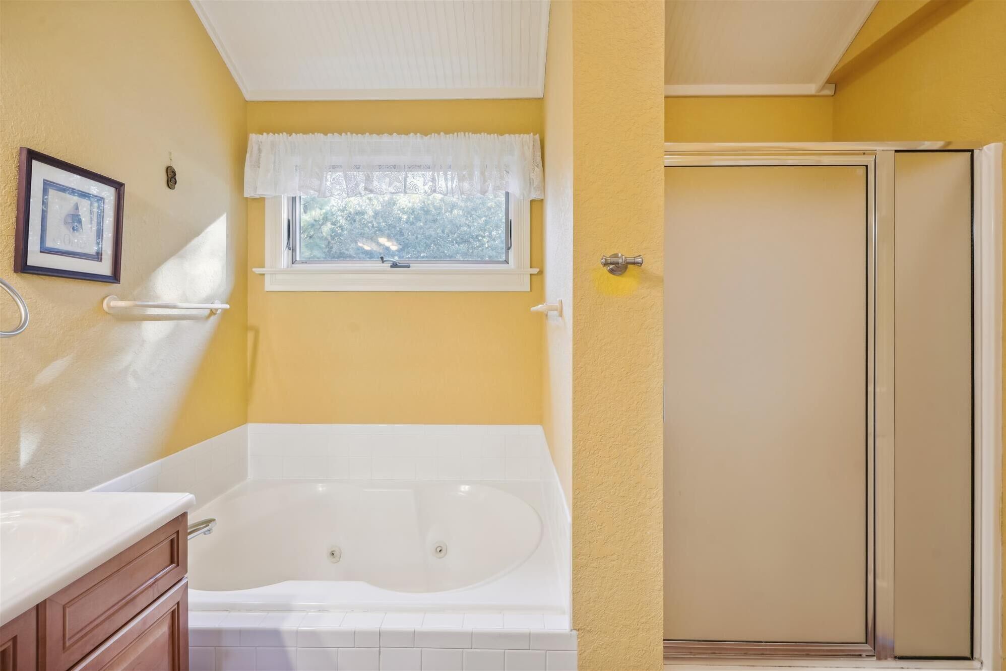 A bathroom with a bathtub, a window with a lace curtain, a framed picture on the wall, and a door with a towel rack.