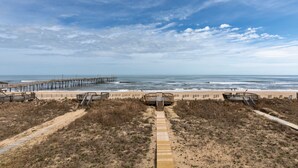 Une plage avec une promenade en bois menant à une structure également en bois, une jetée s’avançant dans l’océan et un ciel dégagé.