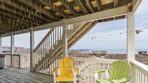 Une terrasse en bois avec deux fauteuils Adirondack, une vue sur la plage et une jetée en arrière-plan.