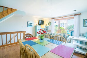 A dining area with a wooden table, chairs, a colorful placemat, and a vase with red flowers. A television is mounted on the wall, and there is a window with curtains.