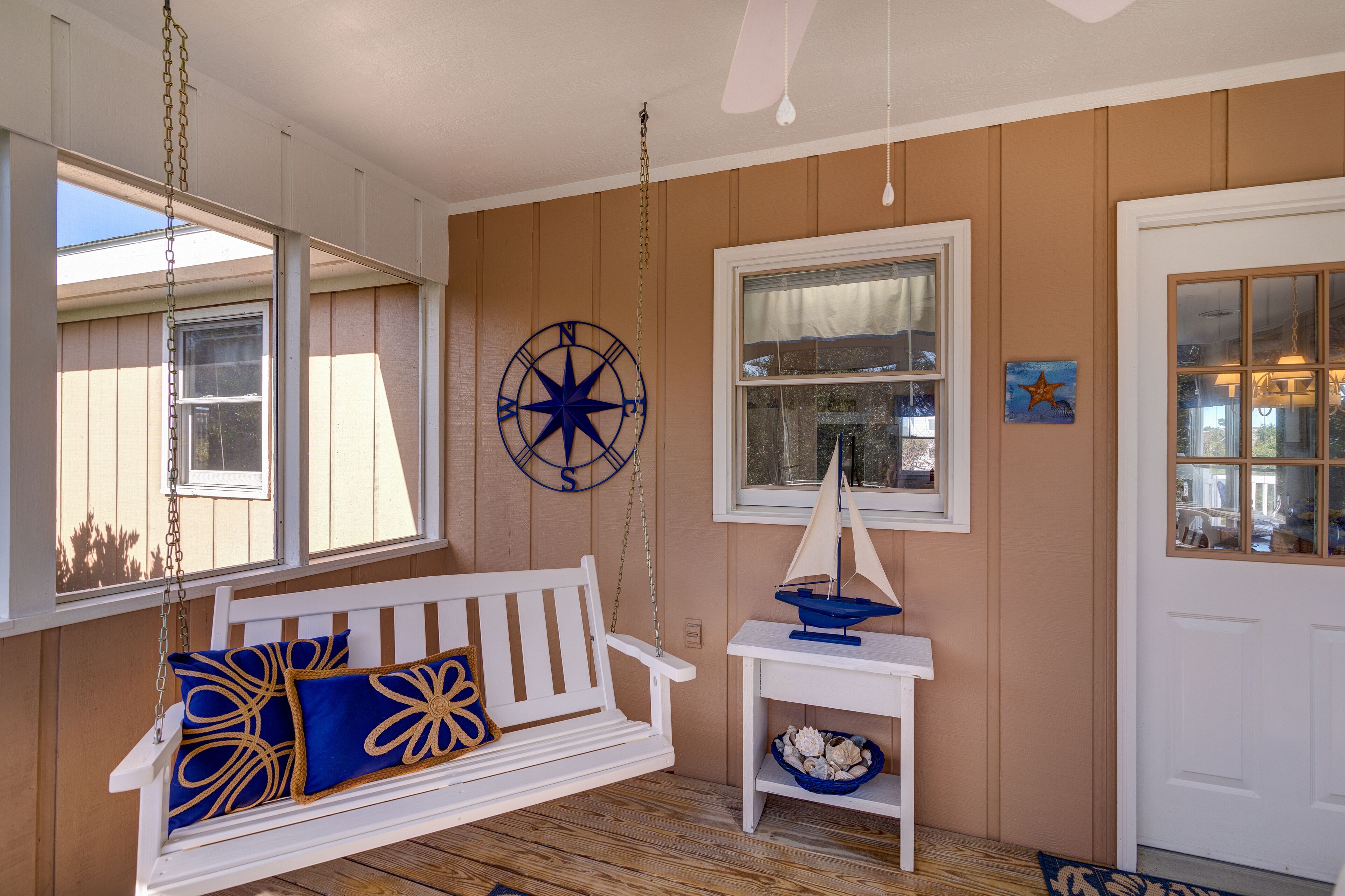 A cozy porch with a white swing, a small table with a sailboat model, and a compass wall decoration.