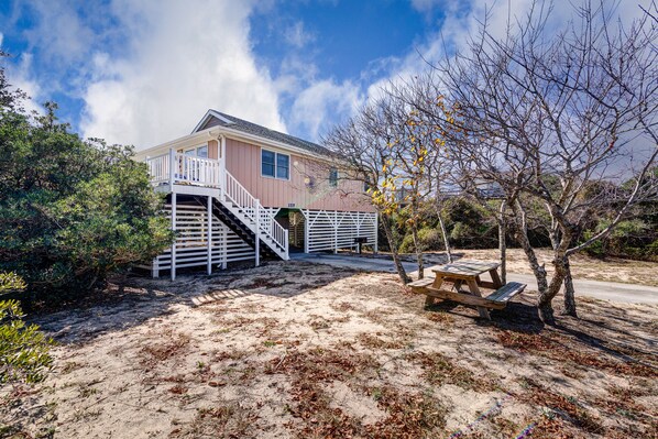 A two-story house with a balcony, a staircase, and a picnic table in a sandy area with sparse vegetation.