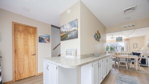 A kitchen with a central island, wooden door, and a dining area with chairs and a table. - 4218- The Scotts' SandBar - Oceanside Duck Condo Near Beach & Dining (Duck)