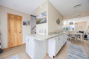 A kitchen with a central island, wooden door, and a dining area with chairs and a table. - 4218- The Scotts' SandBar - Oceanside Duck Condo Near Beach & Dining (Duck)