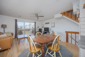 A spacious living area with a wooden dining table, comfortable seating, a television, and a balcony with a view.