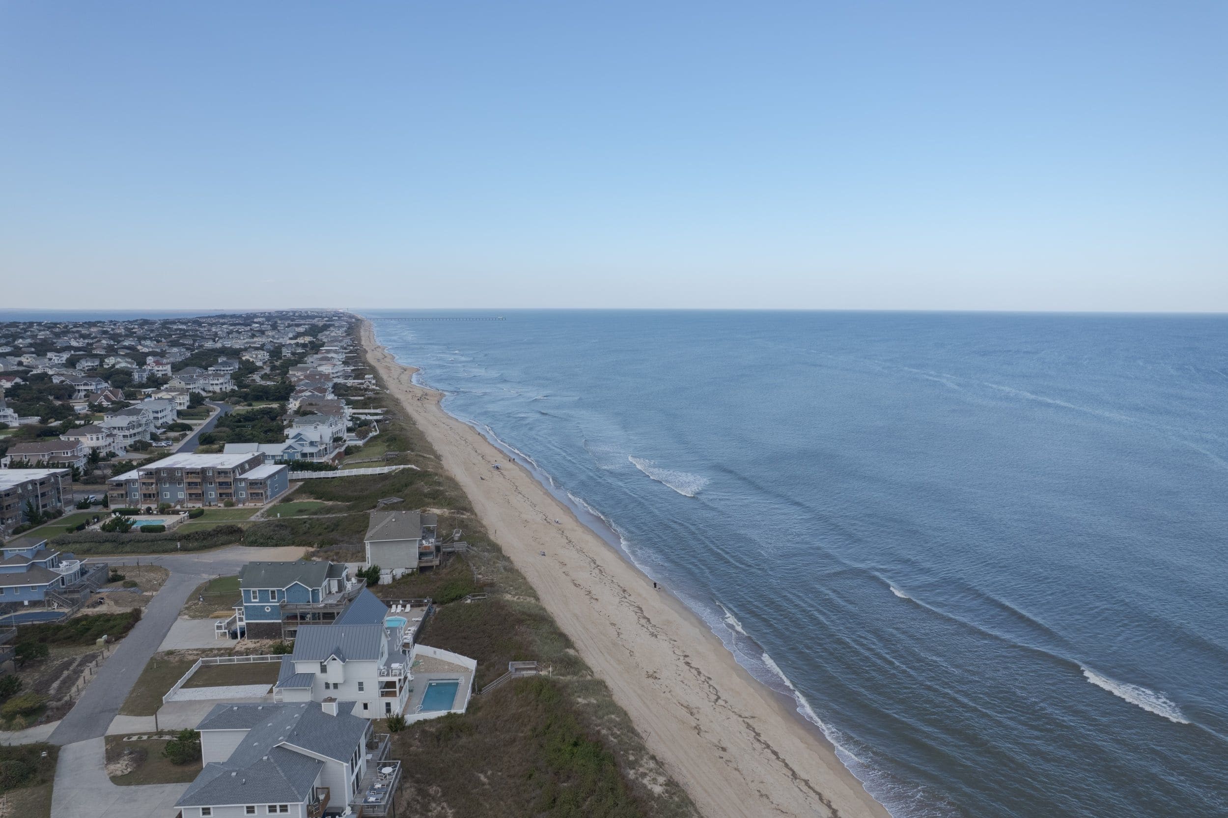 A coastal residential area with houses along the beach, a pier extending into the ocean, and a clear sky.
