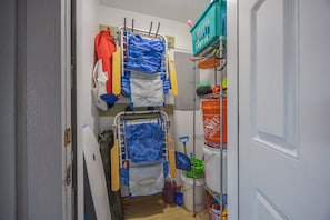 A narrow hallway with a metal drying rack holding various items, a white door, and a shelf with cleaning supplies.