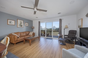 A spacious living room with a ceiling fan, a large window, a sliding glass door leading to an outdoor deck, a leather sofa, a glass coffee table, a TV, and a modern armchair.