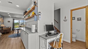 Una cocina moderna con armarios blancos, estantes de madera y una isla integrada con un fregadero. Hay una zona de comedor con una mesa y sillas, además de un escritorio con una silla.