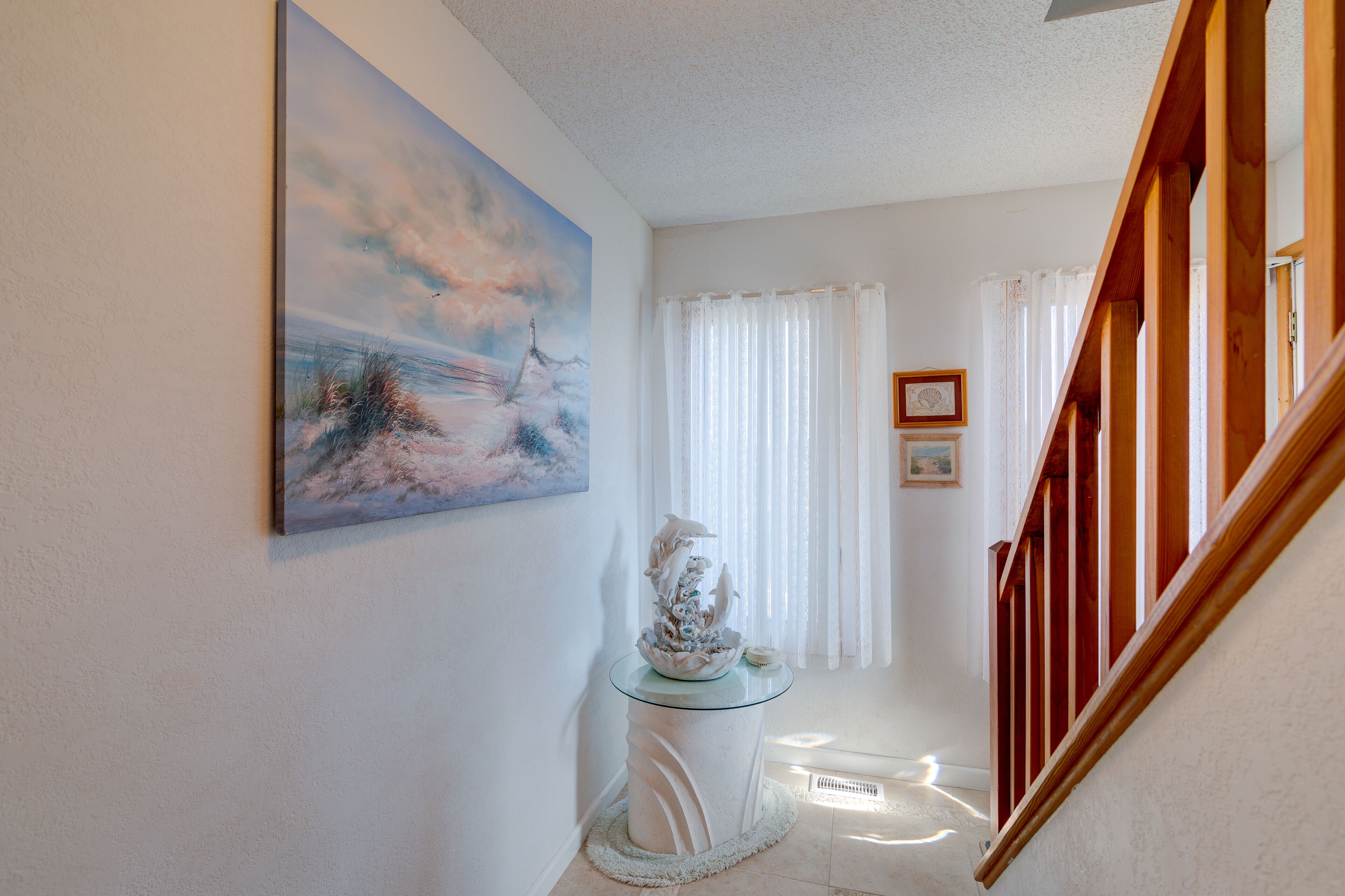 A hallway with a painting of a beach scene, a glass table with a decorative item, and a staircase with wooden railings.
