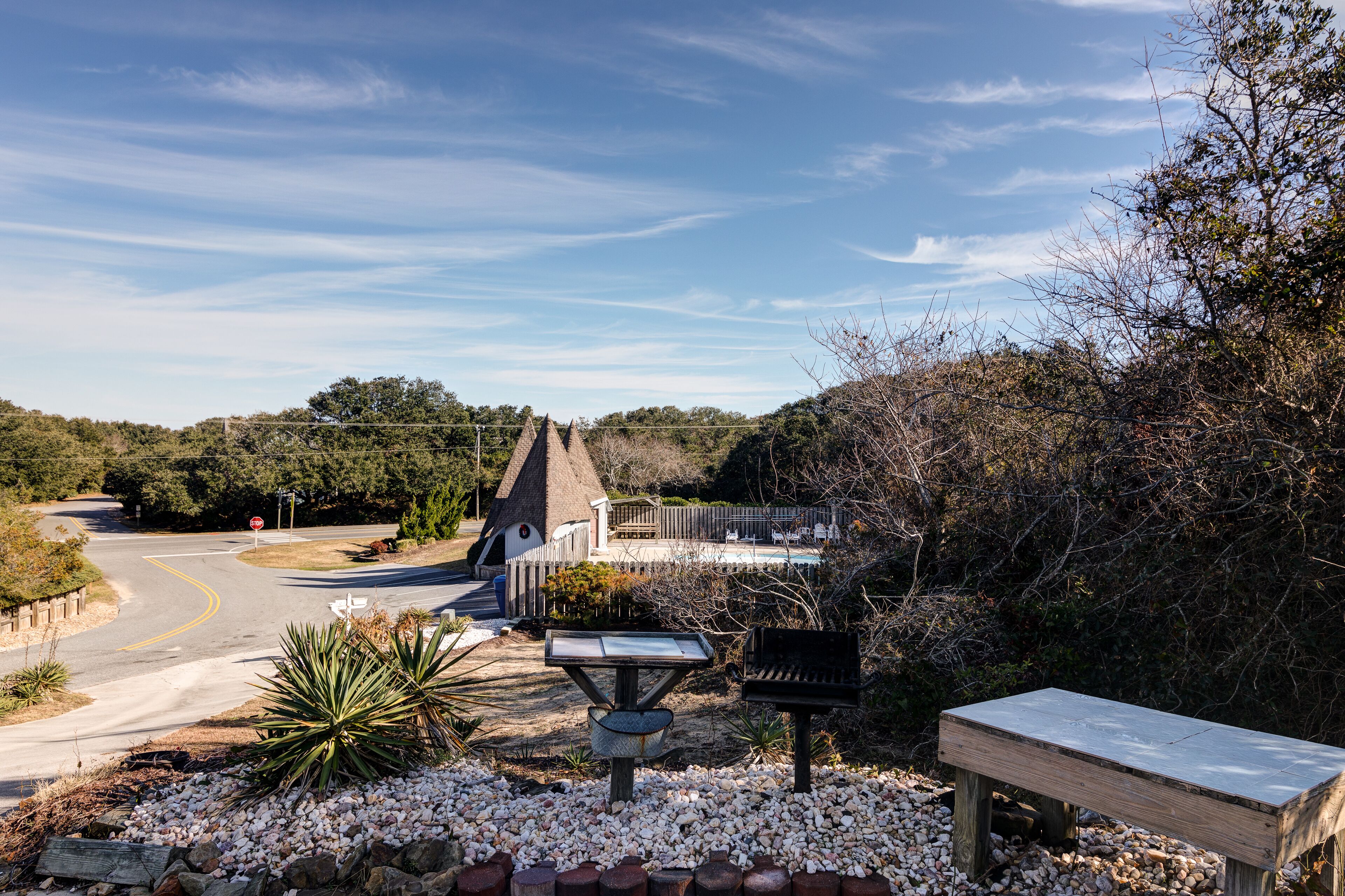 A road curving to the left with a stop sign, a wooden bench, a solar-powered information kiosk, and a thatched-roof structure in a landscaped area.