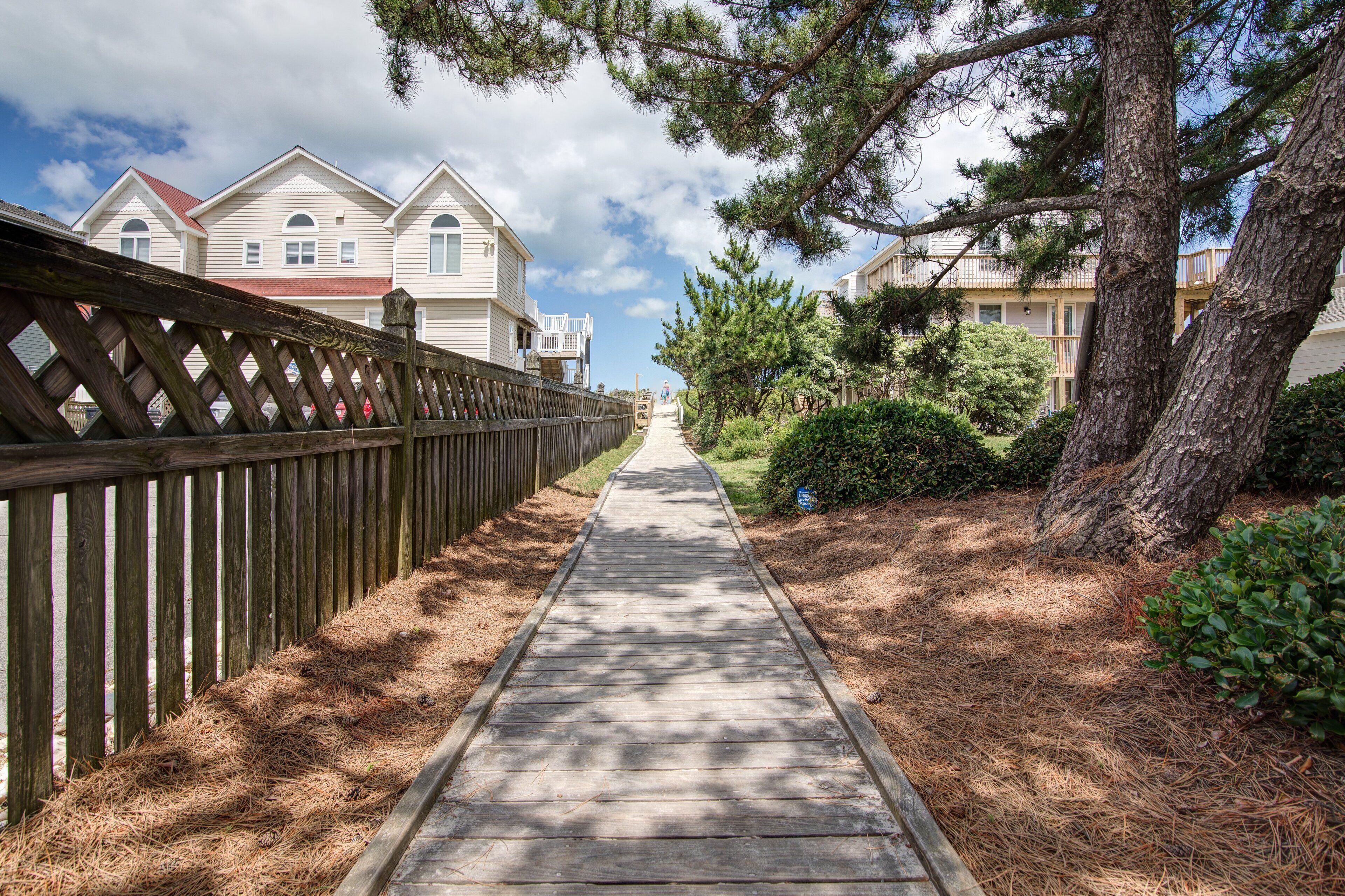A wooden pathway leading to a row of two-story houses with a red roof, surrounded by pine trees and a wooden fence.
