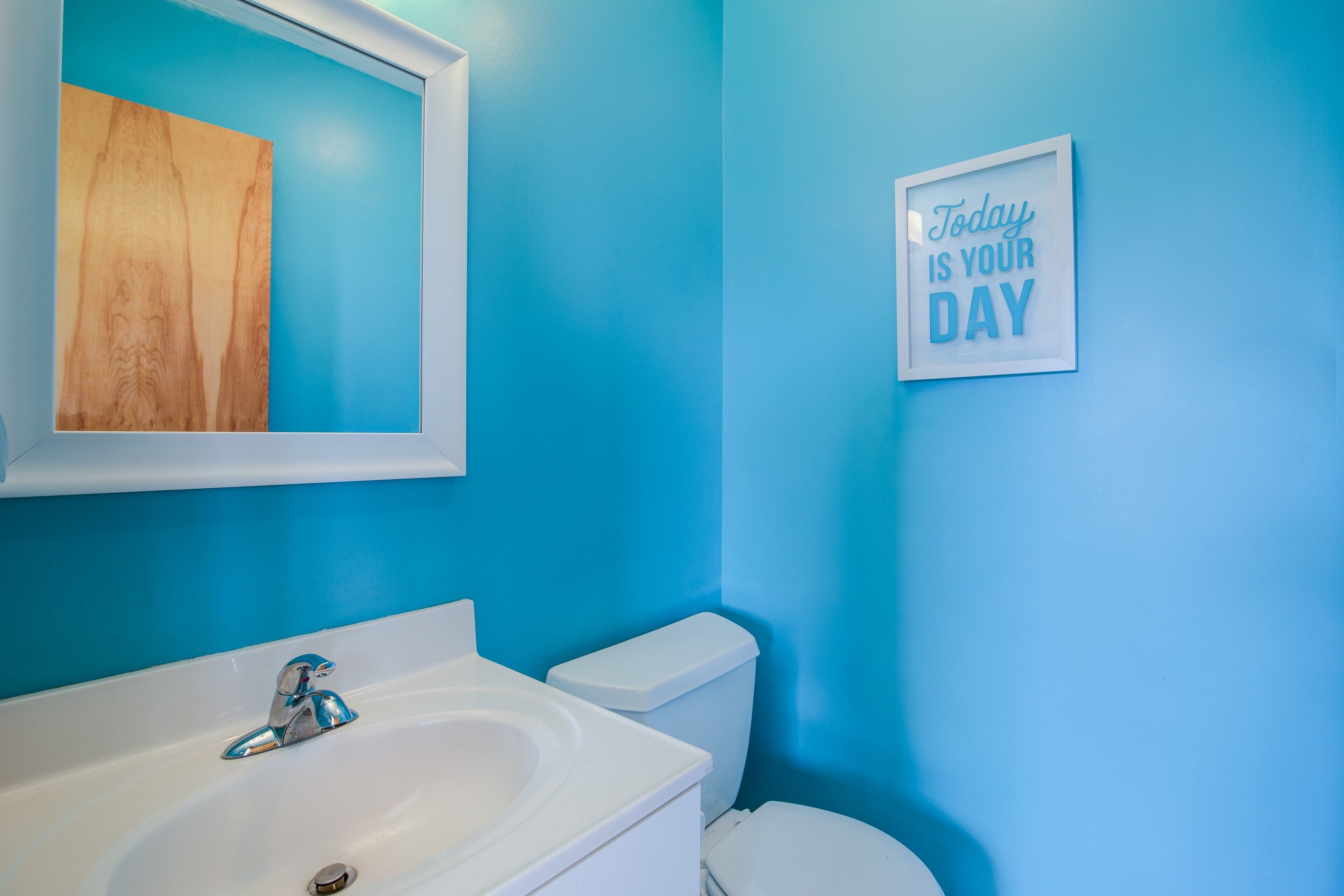 Une salle de bain avec un mur bleu, un lavabo blanc et des toilettes. Un cadre au mur porte une citation : « Aujourd'hui, c'est votre jour. »