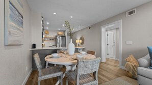 A dining area with a round table, wicker chairs, and a white vase with branches. - 5619- Pura Vida, Oceanfront fully equiped condo! (Nags Head)