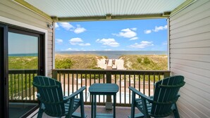 A covered balcony with two Adirondack chairs and a small table, overlooking a beach and ocean. - 5619- Pura Vida, Oceanfront fully equiped condo! (Nags Head)