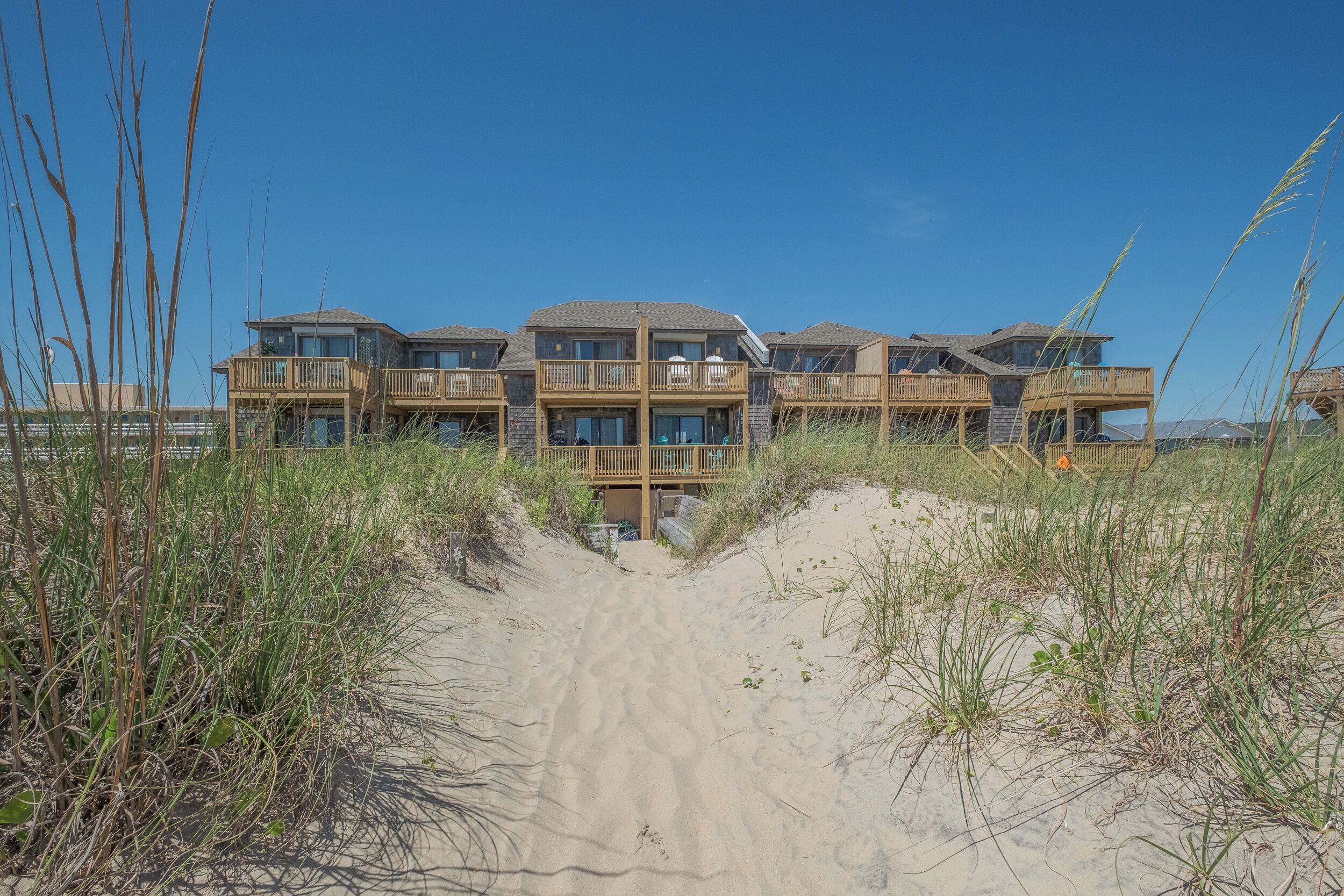 A beachfront hotel with multiple balconies, a sandy pathway leading to the entrance, and surrounding tall grasses.