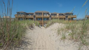 Un hotel frente a la playa con varios balcones, un sendero de arena que lleva a la entrada y hierbas altas que lo rodean.