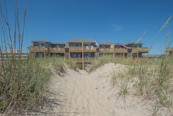 A beachfront hotel with multiple balconies, a sandy pathway leading to the entrance, and surrounding tall grasses.