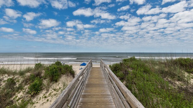 Een houten looppad dat naar een strand leidt, met een heldere lucht en enkele wolken.