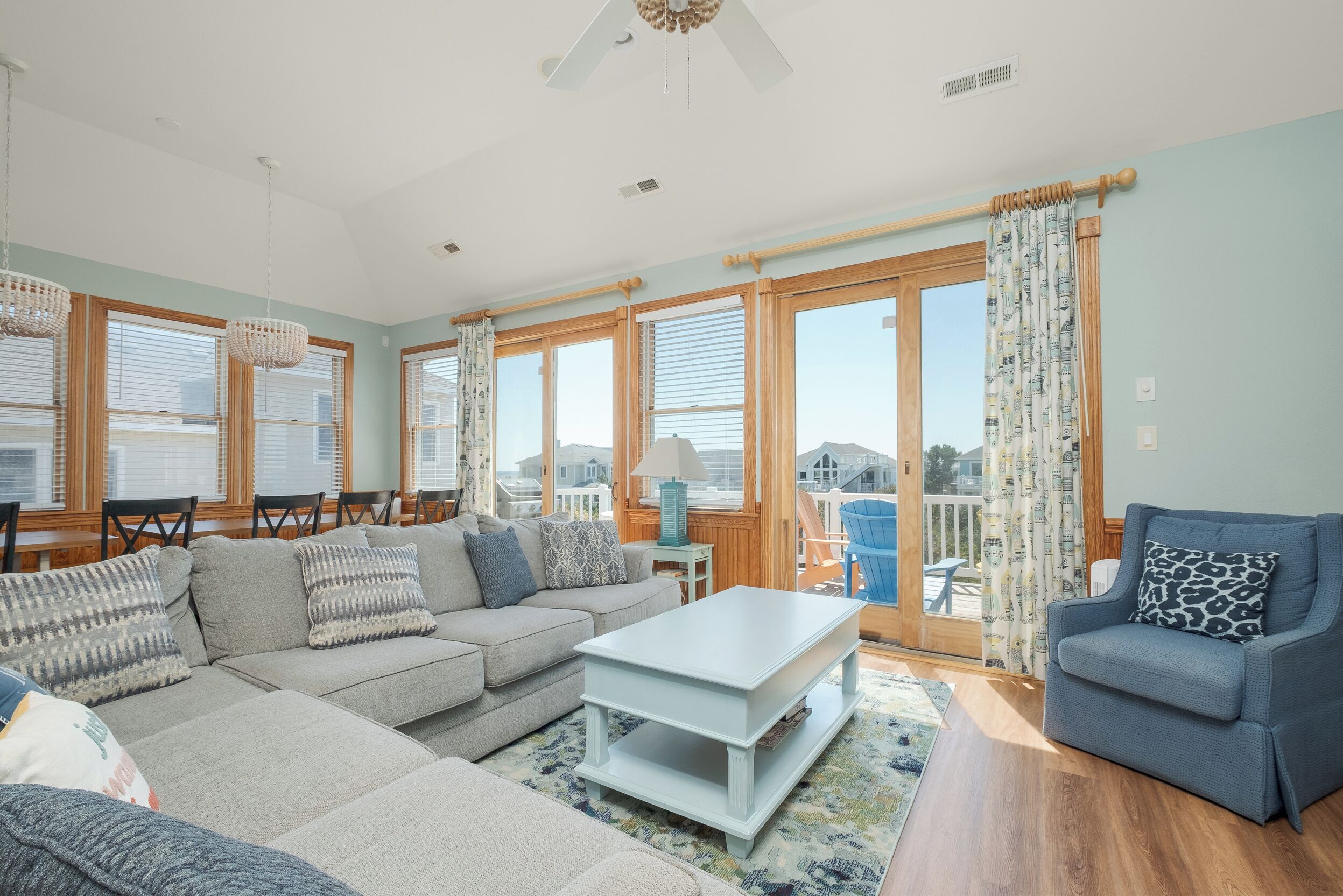 A spacious living room with a large sofa, a coffee table, and a blue armchair. There are wooden-framed windows and a view of the outdoors.