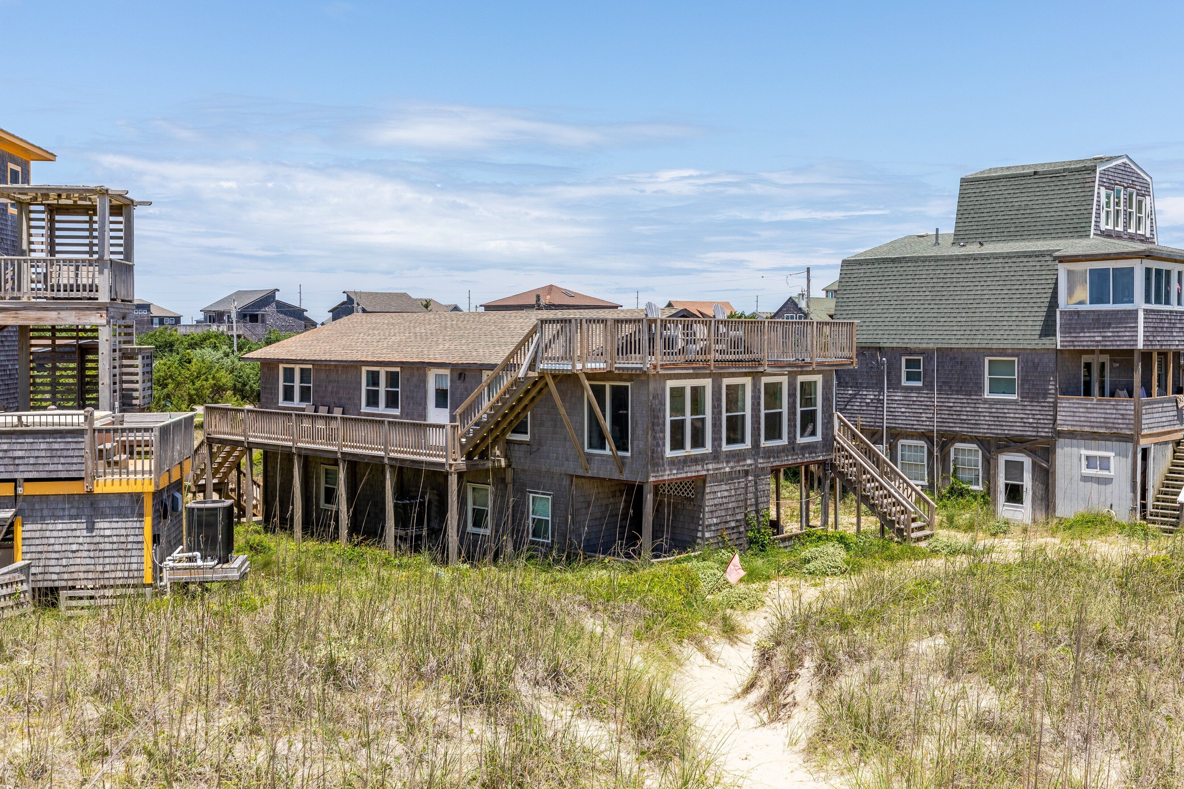 Een huis aan de kust met een houten terras en meerdere verdiepingen, omgeven door duinen en andere huizen.