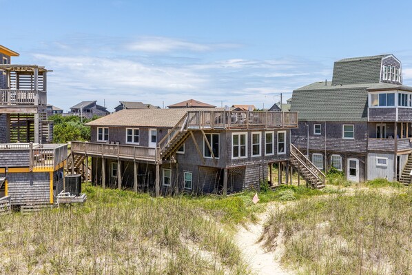 A coastal house with a wooden deck and multiple levels, surrounded by sand dunes and other houses.