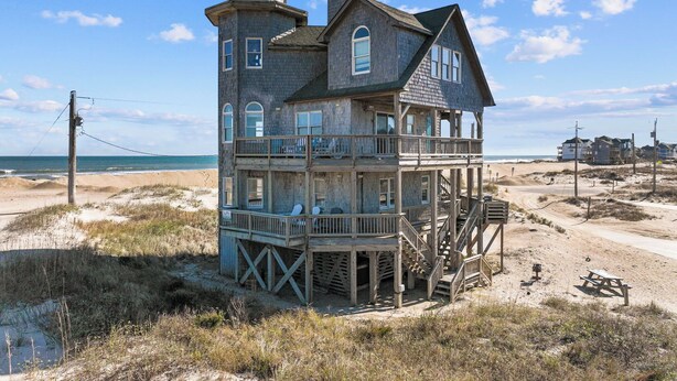 Une grande maison en bois à plusieurs étages, dotée d’un balcon, située sur une plage offrant une vue dégagée sur l’océan.