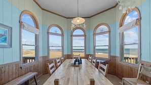 Une table à manger en bois, des chaises, de grandes fenêtres en arc de cercle offrant une vue sur la plage et un luminaire suspendu.