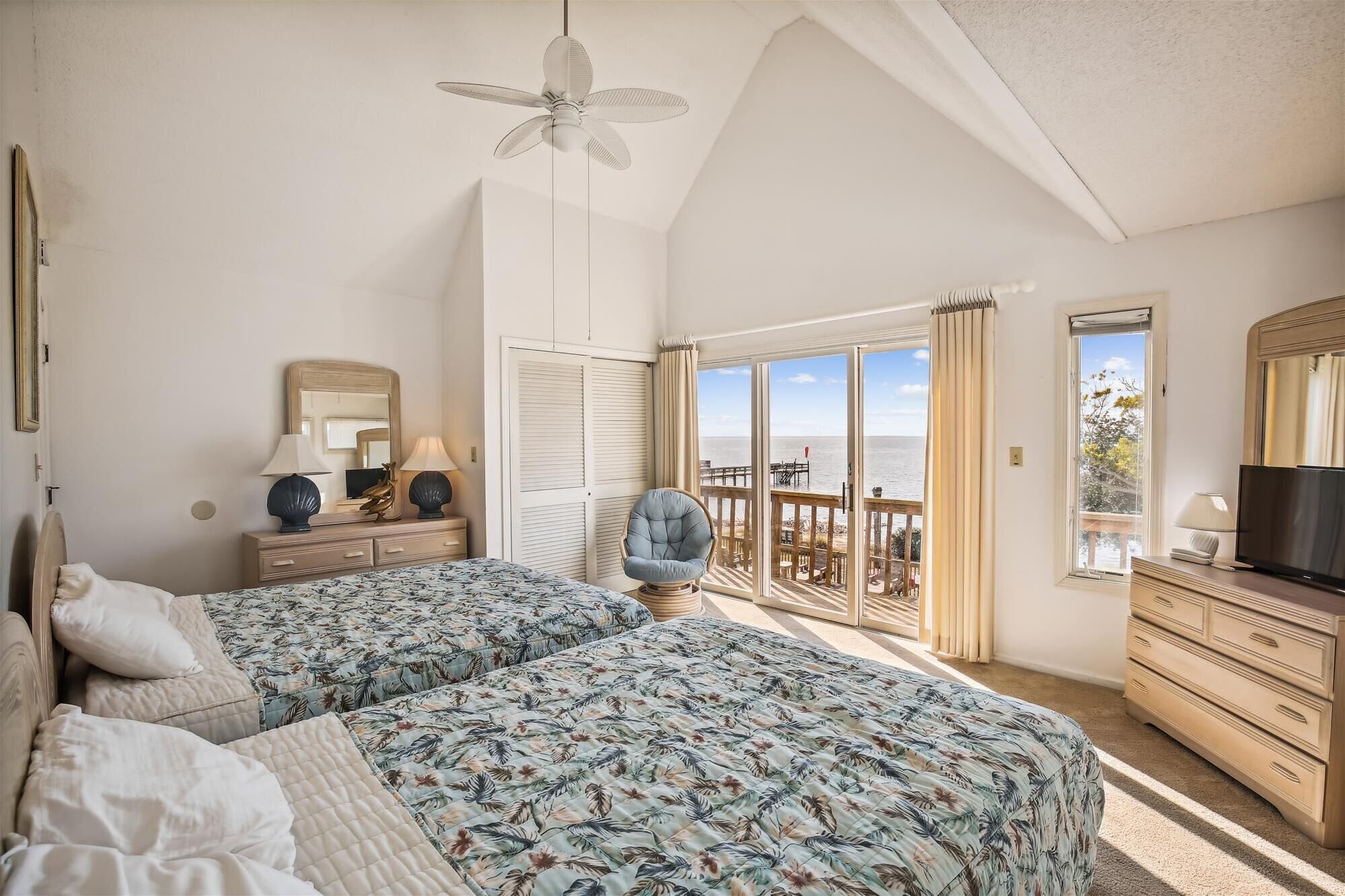 Ein Schlafzimmer mit zwei Betten, einem Deckenventilator, einem Schminktisch, einem Fernseher und einem Balkon mit Meerblick.