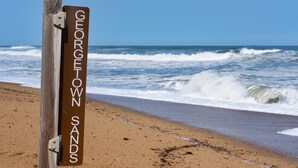Uma placa de madeira com a inscrição 'GEORGETOWN SANDS', colocada numa praia com ondas ao fundo.
