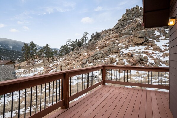 A wooden deck with a metal railing overlooking a rocky hillside with sparse trees and a mountain in the background.