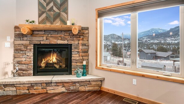 Ein Kamin aus Stein mit Feuer, ein Holzbalken darüber und Blick auf verschneite Berge durch ein großes Fenster.