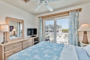 A bedroom with a bed, dresser, TV, and a balcony with a view of houses. - 1886 - Got Beach - Corolla Semi-Oceanfront | Pool & Hot Tub (Corolla)